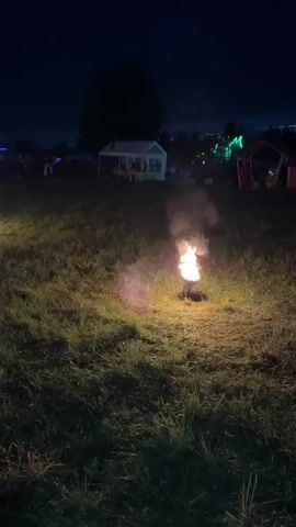 The video captures a large gathering of people at night in an outdoor setting, possibly a festival or concert. People are seen holding up phones to record the event, and there are several small fires burning in the foreground. The sky is dark, indicating that it is nighttime.