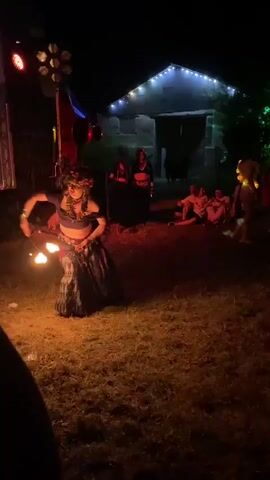 A woman in a dark outfit with a patterned skirt and a flower crown is performing a fire dance outdoors at night. She is holding two flaming poi, one in each hand, and spinning them. In the background, there is a building with lights strung across its roof, and several people are visible, some sitting and some standing. There is also a stage-like structure with lights and a screen to the left. The ground is covered in dry grass and some scattered litter. The overall atmosphere suggests an outdoor event or festival.