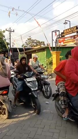 The video shows a street scene with several people riding motorcycles. In the foreground, a person wearing a brown hooded sweatshirt and blue shorts is riding a black scooter. Behind them, an older person wearing a white cap and a light blue shirt is riding a black motorcycle. Further back, a group of people on motorcycles are visible, some wearing hijabs. In the background, there is a green building with signage that includes the words "MAKAM JAMBANGAN TIMUR" and "REPUBLIK INDONESIA". There are also power lines and trees visible. The motorcycles appear to be in a queue, suggesting a traffic jam or a gathering. The overall environment seems to be an outdoor, possibly urban or semi-urban area, with a bright sky and sunlight.