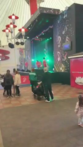 The video shows a group of people watching a performance on a stage. The stage is decorated with musical notes and lights. The performers are wearing red outfits, and one of them is using a hula hoop. The audience includes families with strollers. The environment appears to be an indoor event space or amusement park, indicated by the tent-like structure overhead and decorative elements.