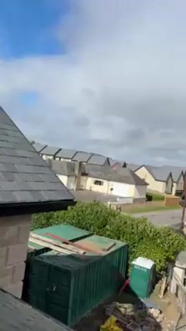 The video shows a view from a height, possibly a rooftop, looking out over a neighborhood of houses under a cloudy sky with patches of blue. The houses have gray roofs and varying facades. In the foreground, on the same level as the viewpoint, there's a section of a gray-tiled roof and part of a stone wall. Below, there are green storage containers and a portable toilet. A road runs through the neighborhood, and a car can be seen driving on it. The general atmosphere is overcast.