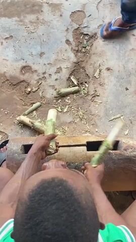 An overhead shot captures several individuals engaged in a musical performance or practice outdoors on a dirt or concrete surface. Two large, weathered wooden logs, each featuring a rectangular opening, are laid horizontally on the ground, serving as percussion instruments (likely slit drums or log drums).

One individual, seen from the back, wears a green and white striped shirt, black shorts, and black flip-flops. They are actively striking one of the wooden logs with a stick in each hand. Another person, partially visible from the chest down, wears brown pants and flip-flops, also striking a log with sticks. A third person's hands are seen holding and striking a round, flat wooden object, possibly a hand drum.

Small pieces of wood and plant debris are scattered across the ground. The scene suggests a traditional music-making session, characterized by rhythmic drumming on wooden instruments in a rustic, outdoor setting. The activity takes place during the daytime, and the location is Ogbu, Nigeria.