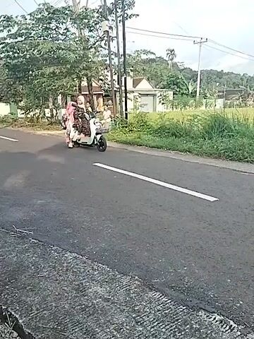 The video captures a serene, overcast daytime scene in a rural area of Bangsri, Indonesia. It begins by showing a woman, wearing a patterned hijab and dress, riding a light green scooter. Two young girls, dressed in pink hijabs and dresses, are seated behind her on the scooter. They are traveling on a paved road marked with a single white line. In the background, other children are visible near residential houses, one riding a bicycle.

As the scooter passes, the camera pans to reveal expansive green fields, likely rice paddies, stretching alongside the road. Utility poles and distant hills frame the landscape. The perspective then shifts to that of a motorcycle rider, with a rearview mirror reflecting nearby trees. Ahead on the road, another motorcyclist, wearing a red jacket and helmet, is seen, followed by a more distant scooter or small motorcycle. The road ahead features a broken white center line. Further along, an arched structure decorated with red and white Indonesian flags marks what appears to be a village entrance. A motorcycle license plate "AD 5899 GN" is partially visible, indicating a vehicle from Central Java. The overall activity depicts daily commuting and life in a peaceful, agricultural community.