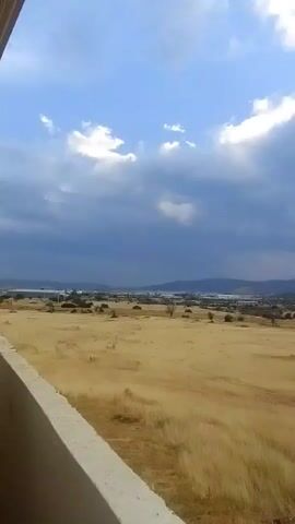 The video shows a wide landscape with a dry, grassy field in the foreground and hills in the distance under a cloudy sky. A structure, possibly a balcony or wall, is visible on the left side of the frame. The sun is visible through breaks in the clouds, casting rays of light onto the scene. The overall impression is of a natural, open environment.