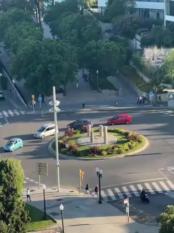 The video presents an elevated, static view of a vibrant urban roundabout and its surroundings during what appears to be a sunny day, possibly late afternoon or early morning due to the quality of light and shadows.

At the center of the frame is a well-maintained roundabout, featuring a circular island adorned with lush green plants, small bushes, and three light-colored, cylindrical decorative structures. Several vehicles are actively navigating the roundabout: a red car, a dark grey SUV, and a silver minivan are seen driving around the central island, while a light blue compact car enters from the left.

Pedestrian activity is also visible. People are seen walking on the sidewalks and utilizing the marked crosswalks. In the lower middle section, two individuals cross a street, and another person walks along the left sidewalk. On the lower right, a person on a black scooter or moped passes through the scene.

The streets surrounding the roundabout are clearly marked with white crosswalks and include traffic lights and streetlights. A blue traffic sign with a 'P' for parking and an arrow is discernible on the upper left, pointing towards a parking area. In the background, the area is characterized by abundant green trees, lining the roads and providing a verdant backdrop. Modern buildings are partially visible further back, integrated into the green landscape. The overall scene depicts a typical, busy urban intersection with both vehicular and pedestrian traffic under clear, bright weather conditions.