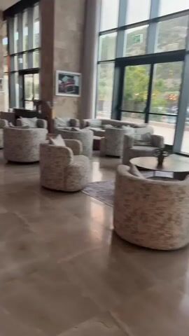 The video pans through a spacious, modern hotel lobby. The flooring is a polished, light-colored marble with a subtle veining pattern. The walls are a mix of textured stone and a wooden slatted design. Large windows offer a view of greenery outside, with the lighting suggesting it's daytime.

In the foreground, several plush, swivel armchairs with a textured, greyish-white upholstery are arranged in small seating groups. Some have scattered cushions. In the mid-ground, more seating areas are visible, including larger, modular white sofas and more of the swivel armchairs.

On the left side of the video, a man in a white t-shirt with a graphic print and dark shorts walks away from the camera. Further back on the left, there are more seating areas and a counter or desk.

In the background, a wall features a striking green, textured design, possibly moss or a similar natural material, interspersed with wooden shelves displaying decor and objects. Hanging pendant lights with woven shades illuminate this area. A bar or cafe setup is also visible in the background with people present. The overall atmosphere is one of contemporary comfort and spaciousness.