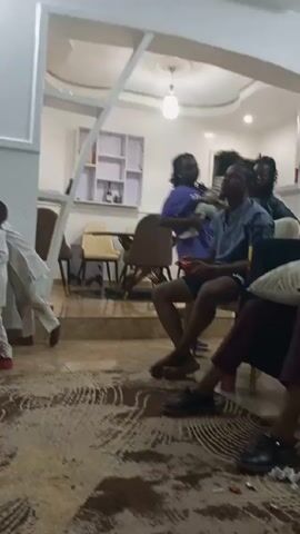 The video appears to be taken indoors in a living room. Several people are present, some sitting and others standing. A man in a blue shirt and shorts is sitting in a chair looking upwards. Two women and a child are standing in the background. Another man in a white robe walks across the room. The video then pans to the left, showing people sitting on a couch. There is a TV mounted on the wall playing a video with nature scenes. The room has a patterned rug and tiled floor. The lighting is provided by ceiling lights.
