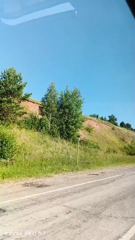The video shows a view from a moving vehicle, likely a car, looking out the side window. The scene is a rural landscape with a clear blue sky. To the left, a grassy slope with scattered trees and vegetation rises up. The slope has exposed reddish-brown earth in patches, indicating erosion or natural soil layers. A road, appearing to be asphalt with some wear and tear, runs along the bottom of the frame. The vehicle's window reflects the sky. The overall impression is of a sunny day in a natural, undeveloped area.