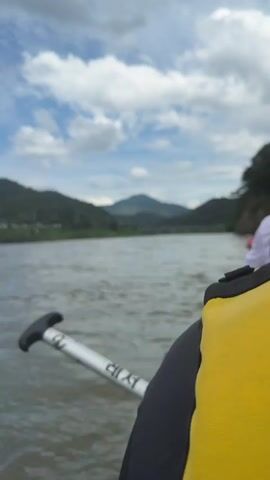 The video shows a first-person perspective of someone in a raft on a river. A yellow life vest is visible in the foreground. A paddle with Korean text "레저" is in the water to the left. In the distance, there are green hills and a cloudy sky. The raft is moving forward, and the water is rippling. At 00:04, another raft filled with people wearing yellow life vests is visible ahead. The other raft is red and appears to be part of a group activity like rafting.