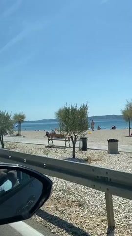 A video shows a beach scene through a car window. The beach has sand and pebbles, with scattered umbrellas and beach chairs. Several people are visible on the beach, some relaxing and others walking. In the background, the calm sea stretches to the horizon under a clear blue sky. A sign for "Konoba Anton Grill" is visible on the right side of the screen. The car appears to be moving along a road adjacent to the beach, with a metal barrier separating the road from the beach area.