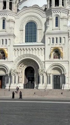 The video shows the facade of a large, white building with ornate architectural details, including multiple domes, arched windows, and decorative carvings. Three people are visible in front of the building: two standing near the entrance and one in a wheelchair. The person in the wheelchair appears to be moving away from the building. The ground in front of the building is paved. The sky is blue and clear. The building is likely a cathedral or a similar religious or historical structure.