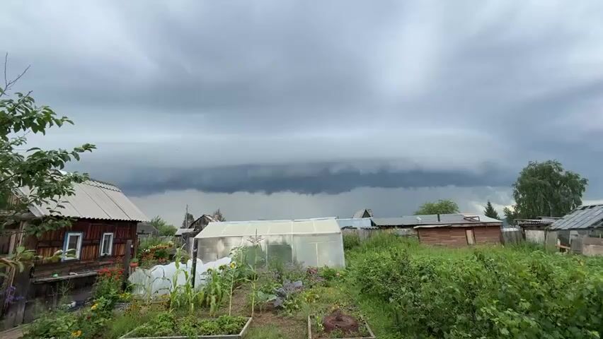 A wide shot shows a village landscape under a dramatic, dark, and ominous sky. The sky is filled with heavy, layered clouds, with a distinct, thick, dark band stretching horizontally across the frame, suggesting an approaching storm. In the foreground, a small garden plot is visible with various plants, including corn, cabbages, and flowers. To the left, a rustic wooden house with a metal roof stands. Adjacent to the garden is a greenhouse made of translucent material. Several other wooden structures and fences are scattered throughout the scene, contributing to the rural atmosphere. Green foliage and trees are present on the right side of the frame. The overall impression is one of anticipation of a significant weather event.