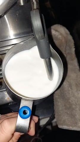 A person's hand, adorned with a ring, holds a metal pitcher filled with white milk. A steam wand is immersed in the milk within the pitcher. The pitcher sits on the surface of an espresso machine, which has a stainless steel finish. Next to the espresso machine, there is a gray towel, partially visible in the background.