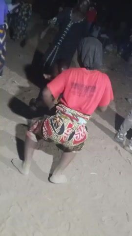 The video captures an individual wearing a red t-shirt, patterned shorts, and a hat, squatting and moving rhythmically in a dusty, outdoor setting. Other people are visible around them, including one in a blue shirt dancing on the ground. The setting appears to be dimly lit and possibly a social gathering, with audible music and dancing contributing to the atmosphere.