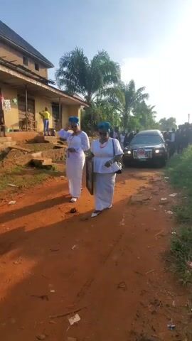 In a medium shot, two women in white dresses and blue head wraps stand on a reddish-brown dirt road. The woman on the left holds her right hand up in a greeting gesture, while the woman on the right carries a framed photo of a man. Behind them, a black car is parked, with several people in dark suits standing around it. In the background, there is a building with a person in a yellow shirt standing on the porch. Palm trees and other vegetation are visible, with a clear blue sky above. The sound of an ambulance siren can be heard in the background.