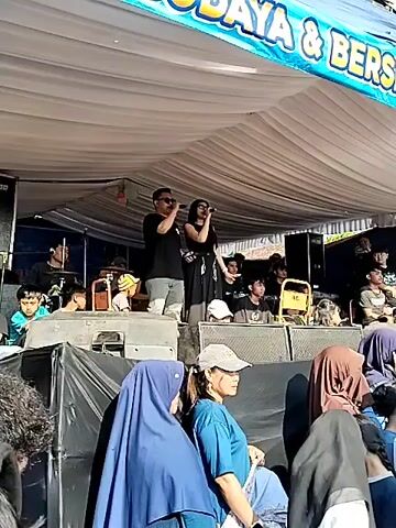 The video shows a live performance on an outdoor stage. Two singers, a man and a woman, are on stage, singing into microphones. The man is wearing a black t-shirt and grey pants, while the woman is wearing a black dress. They are standing in front of a white tent. In the background, there is a banner with text in an unfamiliar language, and some balloons. Below the stage, there are speakers and a crowd of people, some of whom are wearing headscarves. The lighting suggests it is daytime.