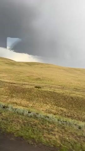 This video captures a panoramic view of a vast, open landscape in Kazakhstan. The scene is dominated by rolling hills covered in dry, golden grass, indicating a dry season or recent harvest. The terrain appears agricultural, with faint tracks suggesting vehicle movement across the fields. In the foreground, patches of green grass and a rough, stony edge of a road are visible.

The sky is dramatic and active, with heavy, dark storm clouds gathering on the left side of the frame, casting a somber mood. As the video progresses, the clouds begin to break apart, allowing shafts of sunlight to pierce through, creating a dynamic contrast between light and shadow. This suggests an impending or passing storm, adding a sense of raw, natural power to the scene. The overall impression is one of solitude and the immense scale of the natural environment. The time of day is not explicitly clear, but the interplay of light and shadow suggests it could be late afternoon or early evening, when storms often intensify. There is no visible text in the video.