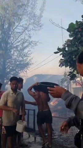 The video shows a group of people reacting to a fire in the background. Several people are carrying buckets, seemingly to assist in putting out the fire, while others are simply watching. The smoke from the fire is visible in the air, obscuring the buildings. A woman in a grey sweatshirt appears distressed, rubbing her eye. The environment seems to be an outdoor setting near a residential area, with trees and power lines visible. The overall tone of the video is one of concern and urgency.