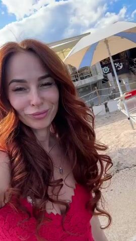 A woman with long, wavy auburn hair is featured in this video. She is wearing a red lace top and appears to be on a beach with the ocean in the background. The sun is shining, and the sky is blue with some clouds. The woman is smiling and looking at the camera. There are buildings and beach chairs visible in the background. The woman walks into the water at one point.