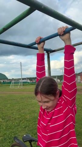 A young woman in a pink and white striped sweater is hanging from a monkey bar set in a grassy field. She is facing forward with a bicycle visible near her on the ground. The sky is cloudy, and houses and a soccer goal can be seen in the background. The woman is using both hands to grip the bar, and then she reaches with one hand to grab the next bar.