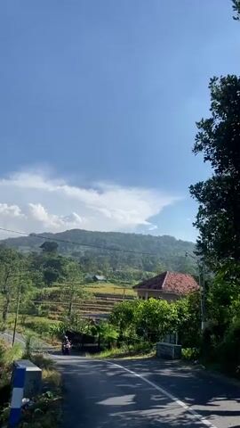 The video shows a scenic view of a rural landscape under a bright blue sky with scattered white clouds. A paved road curves through the scene, with green trees and vegetation lining its sides. In the distance, rolling hills and mountains are visible, some with terraced fields suggesting agriculture. A few houses with terracotta roofs are scattered amongst the greenery. A motorcycle with at least one rider is seen traveling on the road. The overall atmosphere is peaceful and suggests a sunny day in the countryside.