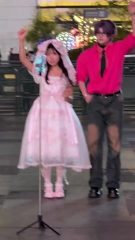 A young woman in a pink and white lolita dress with a headpiece and a young man in a bright pink button-down shirt and distressed gray jeans are dancing together on a tiled walkway outside a building. They are both holding their arms up in the air and moving their bodies to the music. The young woman is standing in front of a phone on a tripod, presumably recording their dance. The young man briefly wraps an arm around her. In the background, there are buildings, decorative lights, and people walking by.
