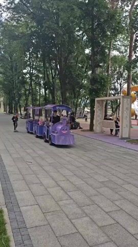 A purple toy train with a smiling face on the front is moving along a paved walkway in a park. The train is pulling two passenger cars, each with a canopy. In the first car, two young children are seated. A young man wearing a black t-shirt and black pants is driving the train. A person on a scooter is visible in the background to the left, and a playground with children is visible in the background to the right. The scene appears to be taking place during the day, with a cloudy sky. The trees are full of green leaves, suggesting it is either spring or summer. The walkway is made of interlocking pavers. There is a bench with people sitting on it to the right of the walkway. Text is visible on the side of the passenger cars and on the front of the train, but it is too blurry to read. The overall mood appears to be lighthearted and cheerful, with families enjoying a day at the park.