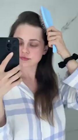 A woman is brushing her hair in front of a mirror. She is wearing a blue and white plaid pajama top and has a black hair tie on her wrist. She is holding a black smartphone in her left hand and using the front-facing camera to record herself. Her right hand is holding a pastel blue and pink hairbrush. The background is a white wall with a marble pattern. She is brushing the right side of her hair down.