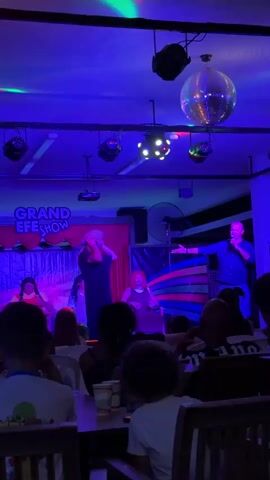 The video shows a stage performance with several people on it. The stage is lit with blue and purple lights, and a disco ball hangs from the ceiling. There is a banner that reads "GRAND EFE SHOW." In the foreground, an audience is seated, watching the performance. On stage, a person with blonde hair in a black dress is in the center, interacting with the audience or another performer. To their left, a person in a red outfit is singing or speaking into a microphone. To the right of the person in the black dress, another person with red hair in a dark dress is also on stage. The person on the far right of the stage is a man in a blue shirt, also holding a microphone and gesturing. The overall atmosphere appears to be a live entertainment event, possibly a drag show or a musical performance.