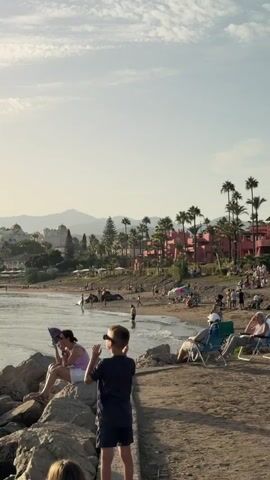 A wide shot of a beach with people enjoying the warm weather. The camera pans from left to right, showing the beachgoers relaxing on lounge chairs, walking along the shore, and interacting with each other. In the background, there are palm trees and buildings, suggesting a tropical or Mediterranean setting. The sun is setting, casting a warm glow over the scene. The video captures a relaxed and peaceful atmosphere.