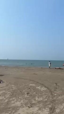 The video shows a beach with a clear blue sky and calm ocean. There are a few people visible: one man standing near the water, one walking across the sand carrying a white plastic bag, and a person driving a motorized cart with a light blue canopy attached. The cart passes from right to left across the frame. There is tire tracks visible in the sand.