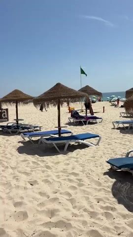 The video shows a sandy beach with several thatched umbrellas and lounge chairs. The umbrellas are spaced out across the sand, providing shade for the chairs. Some of the chairs are empty, while others have people sitting or lying on them. In the background, there are more umbrellas and lounge chairs, and a few people are walking along the shoreline. The sky is blue and clear, suggesting a sunny day. There are two white, pod-like structures on the sand to the right of the frame. A green flag flies from a pole in the middle distance.