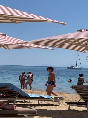 The video shows a beach scene with several people. In the foreground, two beach chairs with striped cushions are visible on the sand, partially obscuring the view. Two large, striped umbrellas are prominent in the upper portion of the frame. In the middle ground, a woman in blue shorts and a dark top walks towards the water. Further back, two women in bikinis stand in the shallow water, facing each other. A sailboat is visible in the background on the blue sea, with a distant landmass on the horizon. Several yellow buoys are scattered in the water. Another woman in a bikini is seen swimming in the water to the right, and a portion of another person on a beach chair is visible in the lower right corner. The sky is clear and blue.