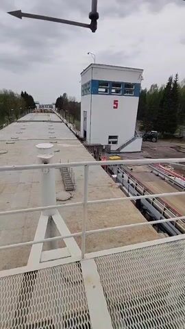 The view is from a high metal platform or bridge, probably at a ship lock. In the distance is a long concrete channel with what appear to be boats in it. A white tower, possibly the control center for the lock, is visible with a large red number "5" on its side. The tower has blue trim around the top and what appear to be windows. A staircase leads up the side of the tower. To the right of the tower is a tractor, and beyond are trees. A weather vane is visible at the top of the frame, with the arrow pointing left. The sky is overcast.