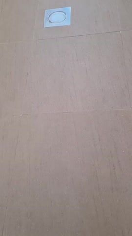 This is a close-up, low-angle shot of a tiled floor. The tiles are large and square, in a light gray color with a subtle texture. There are thin, dark grout lines between the tiles. In the center of the frame, there is a square metal drain cover with a circular opening in the center. A thin, circular object, possibly a piece of wire or string, is visible within the opening of the drain cover. The video is static and does not show any movement or activity.