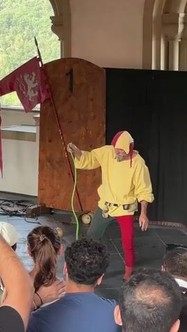A man dressed as a jester, wearing a yellow tunic with red and yellow headwear, red pants, and green leggings, performs a juggling act with a green rope. He is standing on a stage in front of a large wooden door with a number '1' painted on it. A red flag with a white lion emblem is attached to a pole to the left of the door. The stage is set indoors, with stone pillars visible in the background and a black curtain behind the performer. A small audience, mostly seen from behind, watches the performance. There are cables on the floor in front of the stage.