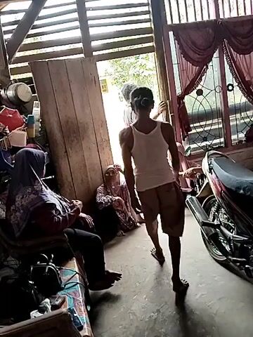 The video shows an indoor scene with several people. In the foreground, a person wearing a hijab is sitting on a chair. To the left of this person, another person in a patterned hijab is sitting on the floor. In the center of the frame, a young man in a white tank top and shorts stands by an open doorway, gesturing with his hand. To his right, another person, possibly a woman, is also sitting on the floor. A motorcycle is visible on the right side of the frame. The environment appears to be a simple dwelling with wooden walls and a corrugated iron roof. The lighting suggests it is daytime, with natural light coming from the open doorway. The people appear to be in conversation or observing the young man.