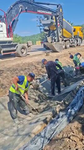A construction crew is pouring concrete into a trench. In the foreground, a man wearing a high-visibility vest and a blue cap shovels concrete into the trench. Behind him, another man in a similar vest is also shoveling. Further back, two more men are working with shovels. To the left, an excavator with the brand name "HIDROMEK" is parked, and a cement truck is positioned behind it, its chute dispensing concrete into the trench. The environment appears to be a construction site with dirt and loose gravel. The sky is blue with some clouds. The video is shot from a slightly elevated angle looking down into the trench.