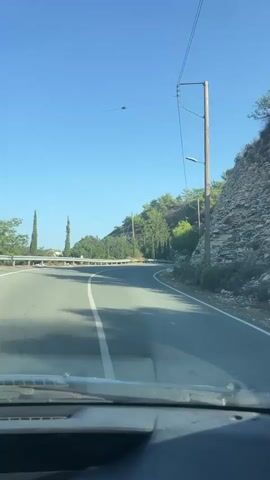 The video shows a view from inside a vehicle traveling on a paved road that curves to the right. The road is bordered by a metal guardrail on the left and a rocky, tree-covered hillside on the right. Tall, slender trees are visible on the left side of the road, and there are power lines running along the right side, supported by wooden poles. The sky is clear and blue. The dashboard and windshield wipers of the vehicle are visible in the foreground.