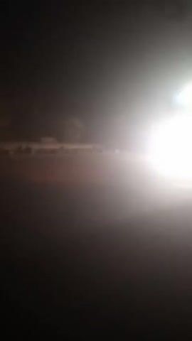The video shows a view from a moving vehicle at night.  The primary focus is the bright headlights of an approaching vehicle, which are creating a glare and obscuring details in the background.  The approaching vehicle appears to be a large truck, possibly a semi-truck, with multiple lights on its roof.  There are other lights visible in the distance, suggesting other vehicles or streetlights. The road surface is dark and appears to be asphalt.  The environment is dark and hazy, possibly due to fog or dust.