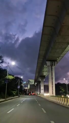 The video shows a road with a metro line overhead at dusk. A road is visible in the foreground with lane markings and streetlights. There are trees and a metro station on the left side of the road, and a metro line supported by concrete pillars on the right. Several vehicles, including cars and motorcycles, are visible on the road. The sky is dark with clouds, and the lights of the vehicles and streetlights illuminate the scene.