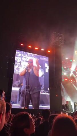 The video shows a concert crowd watching a performance on a large screen. On the screen, a bald man wearing sunglasses and a black tank top and pants is singing into a microphone with his right arm raised. Behind him, the stage is lit up with colorful lights and smoke effects. The words "PARK LIVE" are visible in large illuminated letters on the stage. The crowd in the foreground consists of many people, most of whom are holding up their phones to record the performance. The lighting is dark, typical of a concert venue at night, with bright stage lights illuminating the scene.
