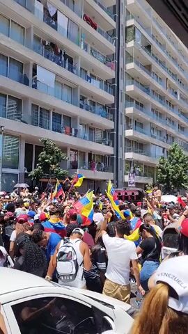 The video shows a large crowd of people celebrating in the street, many of whom are waving Venezuelan flags. They are gathered around a truck with a large sound system. The crowd is dense and appears to be moving forward.  There are apartment buildings in the background, some of whose balconies are occupied by people watching the celebration. The weather appears to be sunny.