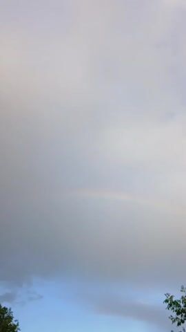 This video captures a cloudy sky with a faint rainbow visible. The lower portion of the video shows glimpses of trees and a brick house. The camera appears to be handheld and is slowly panning upwards.