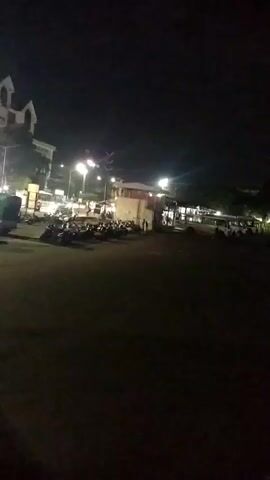 The video depicts a nighttime scene of a parking area for buses. A row of several white buses with various colors and logos sits under a street light, which illuminates them and creates a starburst effect. In the background, there are more parked vehicles and what appear to be buildings with lit windows. The ground is dark and unclear, but seems to be a paved area. The overall lighting is dim, with the street lights being the primary source of illumination. The scene seems quiet and still.