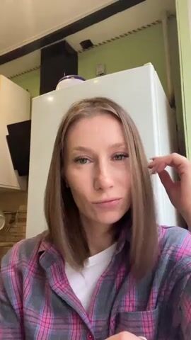 A woman with shoulder-length brown hair and blue eyes is in a kitchen. She is wearing a pink and gray plaid shirt over a white t-shirt. She is touching her hair and looking at the camera with a slight smile. The background shows a white refrigerator and cabinets.