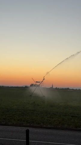A central pivot irrigation system is operating in a field at sunset. The irrigation system is a long metal structure with wheels, and it is spraying water into the air, creating a mist. The sun is setting in the background, casting a warm orange glow over the sky and the field. There is a town or village visible in the distance, with a church steeple. The field appears to be covered in crops. The foreground shows a paved surface, possibly a road, and a dark pole. The video is shot from a low angle.