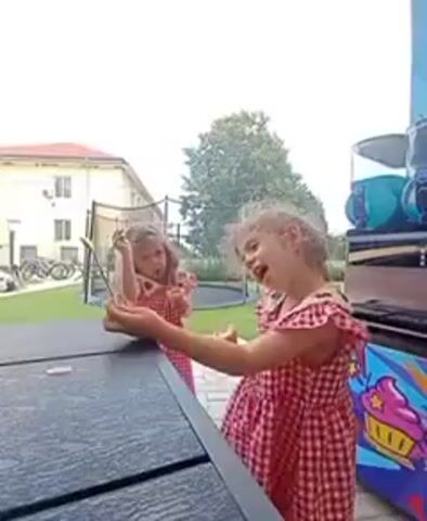 Two young girls in red and white checkered dresses are playing outdoors. One girl, closer to the camera and in the foreground, is laughing and holding a spoon in her right hand. The other girl, further away and in the background, is holding a spoon and appears to be making a gesture. They are near a dark-colored table, and behind them is a trampoline, a building, and trees. A colorful, cartoonish mural featuring a cupcake is visible on the right side of the frame.