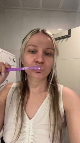 A woman is brushing her teeth in a small, cluttered bathroom. She has blonde hair and is wearing a white tank top and pink pants. She holds a purple toothbrush in her right hand. In the background, there is a washing machine and dryer stacked on top of each other, a white tiled wall, a closed door with a rack, and a ceiling with a square pattern.  Music is playing in the background. The woman alternates brushing her teeth and then touching her face to reposition the toothbrush. The camera is static, with a medium shot focusing on the woman.