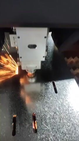 A laser cutting machine is shown in operation. The laser head moves across a sheet of galvanized metal, creating parallel slots. Sparks are visible as the laser cuts through the material. The machine is enclosed and appears to be in a factory or workshop setting. The video is shot from a close-up perspective, focusing on the cutting process.